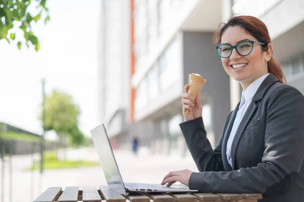 Happy woman travaille sur un ordinateur assis sur une terrasse d'été d'un café et mange de la glace à la vanille. Une employée tape sur un clavier d'ordinateur portable et tient un cône de crème glacée. Dessert rafraîchissant. — Photo