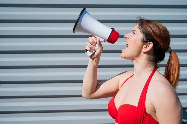 Rothaarige Kaukasierin im roten Bikini, die vor dem Hintergrund einer grauen Wellblechwand durch ein Megafon brüllt. Ein Mädchen im Badeanzug steht mit einem Lautsprecher im Profil. Sommerferien. — Stockfoto