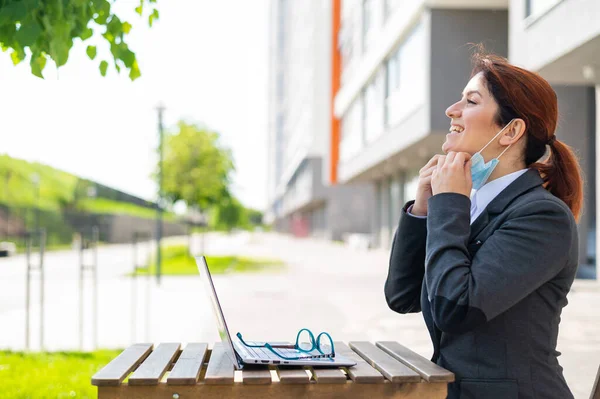 La muchacha feliz en el traje trabaja remotamente en el portátil al aire libre. Una mujer de negocios se quita una máscara médica mientras está sentada en una terraza de verano. Fin de la cuarentena. Coronavirus es derrotado. Salud y libertad —  Fotos de Stock