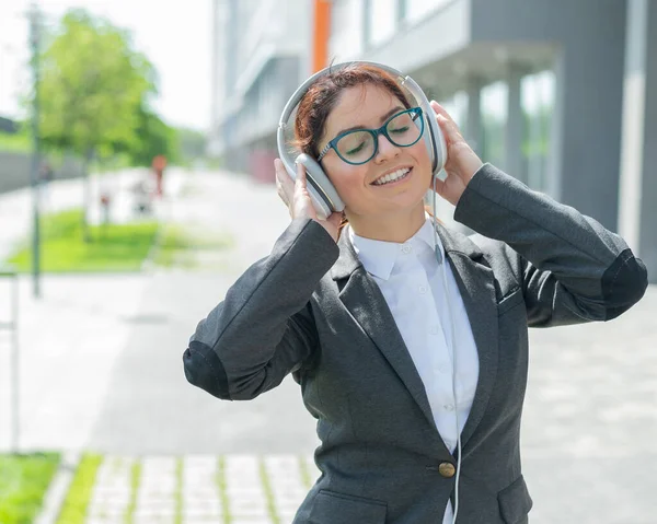 Porträtt av en leende kvinna i kostym som lyssnar på musik i hörlurar utomhus. Kvinnlig kontorsanställd håller händerna på hörlurar njuter sång och dans på gatan. — Stockfoto