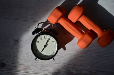 Sabahın köründe spor yapma zamanı. Alarm saatinin üst görüntüsü ve ahşap zeminde turuncu halterler..