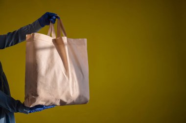 Çevre dostu, sarı arka planda tekrar kullanılabilir pamuk torbası göndermek. Kurye, sarı arka planda plastik ambalajsız bir bez çanta tutuyor. Salgına güvenli bir şekilde teslim.