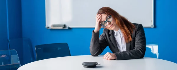 En trøtt forretningskvinne i briller og en dress alene i et tomt møterom. En kontorarbeider med hodepine venter på at presentasjonen skal begynne i styrerommet. – stockfoto