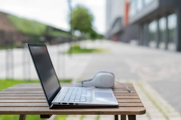 Close-up of a laptop and white big headphones on a wooden table on a summer terrace. No people. Cafe at the outdoors. Freelancer workspace. — Stock Photo, Image