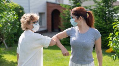 Women in medical masks greet their elbows outside. An elderly woman and her adult daughter maintain a social distance in the park during the coronavirus epidemic. New handshake bumping elbows clipart