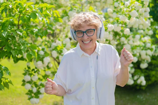 Щаслива стара жінка слухає музику на навушниках і танцює в парку квітучих яблунь. Активна жінка-пенсіонерка посміхається і насолоджується теплим літнім днем у саду. Передове старше покоління . — стокове фото