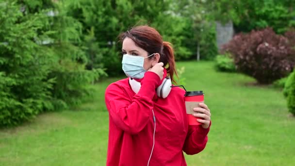 Rudowłosa kobieta w słuchawkach pije kawę ze słomką w parku i zdejmuje maskę medyczną. Dziewczyna korzysta z bezpłatnego spaceru w słoneczny letni dzień pod koniec kwarantanny. — Wideo stockowe