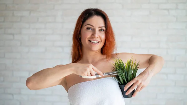 A smiling red-haired woman in a white terry towel cuts a green plant in a pot with scissors. Imitation of armpit depilation. Hair removal concept. Procedure for hair removal of unwanted vegetation.