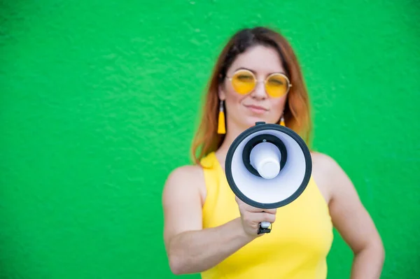 Glückliche Frau in gelbem Kleid mit Brille und Ohrringen auf grünem Hintergrund, die in ein Megafon ruft. Porträt eines Mädchens mit einem Lautsprecher. Dame mit einem perfekten schneeweißen Lächeln. — Stockfoto