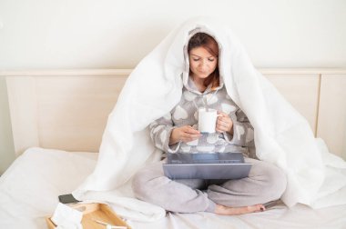Yataktaki kadın kafasıyla battaniyeyle örtülmüş ve sıcaklığı ölçüyor. Soğuk algınlığı olan bir kız dizlerinin üzerinde dizüstü bilgisayarla yatak odasında oturuyor..