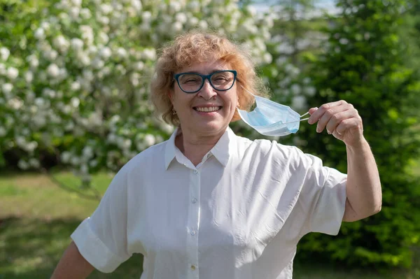 En lycklig äldre kvinna är botad från en allergi mot blomning och tar av sig sin medicinska mask. Mormor njuter av vårblomningen av äppelträd i trädgården. Astma för pollen. — Stockfoto