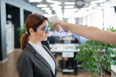 Ofis personeli için elektronik kızılötesi termometre ile sıcaklık ölçümü. Takım elbiseli hasta kadın. Silah termometresinin çalışmasını kontrol etmek yasak. Covid-19 'un yayılmasını önleme.