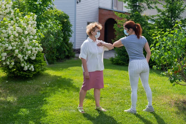Dos mujeres enmascaradas saludan sus codos en el parque. Una mujer anciana y su hija mantienen distancia social durante la epidemia de coronavirus. Una nueva forma de estrechar las manos. Los manzanos están floreciendo . —  Fotos de Stock