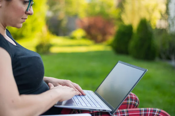 Uma mulher ruiva senta-se em um parque e digita no computador enquanto faz compras on-line. A menina mantém uma distância social e trabalha ao ar livre em um laptop. O estudante está estudando remotamente . — Fotografia de Stock