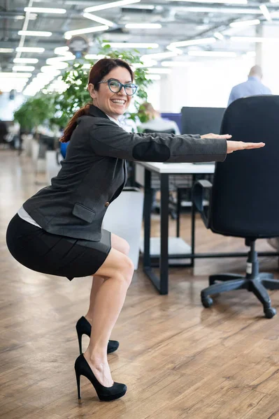 Happy business woman squat in open space office. A red-haired smiling female employee in a skirt and high heels does exercises on the buttocks in the workplace. Sport at work. Fitness as a lifestyle.