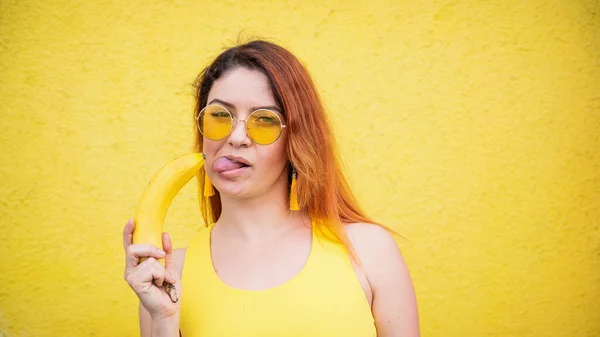 En vacker rödhårig kvinna i gul klänning och solglasögon visar sin tunga och slickar en banan. Porträtt av en fantasifull flicka med mogen frukt. — Stockfoto
