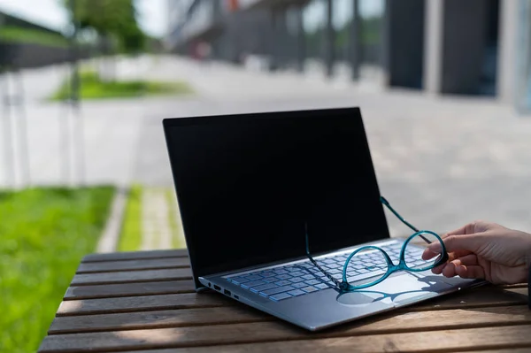 Close-up of a female hand holds glasses near a laptop on a wooden table in a summer cafe. Freelancer workplace. A woman remotely working outdoors. — Stock Photo, Image