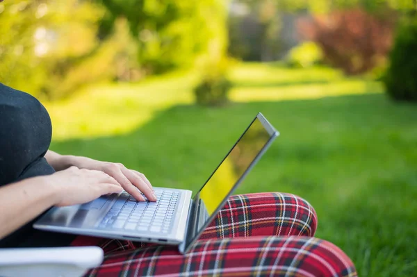 Mulher senta-se em um parque e tipos no computador enquanto faz compras on-line. A menina mantém uma distância social e trabalha ao ar livre em um laptop. O estudante está estudando remotamente . — Fotografia de Stock