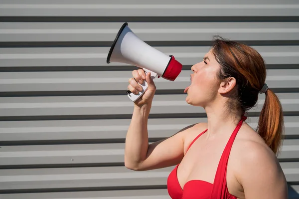 Femme caucasienne aux cheveux roux en bikini rouge criant à travers un mégaphone sur un fond de mur ondulé gris. Une fille en maillot de bain est debout de profil avec un haut-parleur. Vacances d'été. — Photo