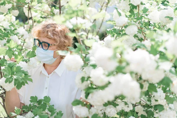Eine ältere Frau mit Brille und medizinischer Maske inmitten der Blüten von Apfelbüschen während der Quarantäne. Eine Allergieattacke während der Blüte im Frühjahr. Großmutter geht während der Isolation im Park spazieren. — Stockfoto