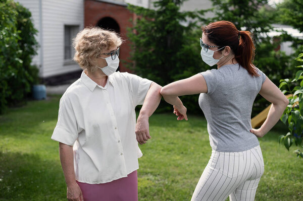 Women in medical masks greet their elbows outside. An elderly woman and her adult daughter maintain a social distance in the park during the coronavirus epidemic. New handshake bumping elbows