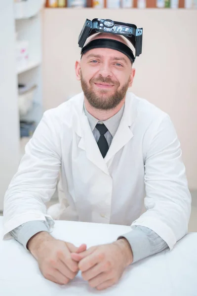 Maschio medico barbuto in un cappotto medico e con una cinghia di ingrandimento w luci seduto sulla scrivania. Attrezzature optometriche. Ufficio medico. — Foto Stock