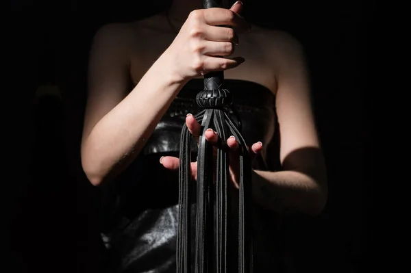 Concept BDSM. Une femme dans un corset en cuir tient un fouet noir avec une longue frange. Jeux de rôle pour adultes. Gros plan sur les jouets sexuels pour la domination. Le fouet dans les mains féminines. — Photo