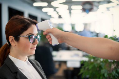 Ofis personeli için elektronik kızılötesi termometre ile sıcaklık ölçümü. Takım elbiseli hasta kadın. Silah termometresinin çalışmasını kontrol etmek yasak. Covid-19 'un yayılmasını önleme.