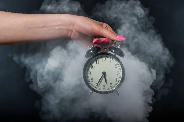 Una mujer sostiene un despertador en un estudio lleno de humo. Niebla blanca envolvió un reloj mecánico retro redondo . —  Fotos de Stock