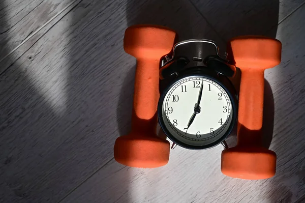 Its time to do sports early in the morning. Top view on alarm clock and orange dumbbells on a wooden floor.