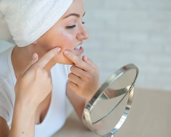 Una mujer con una toalla blanca en la cabeza se mira en el espejo y aprieta un grano en la mejilla. Una chica con problemas hormonales hace limpieza facial en casa. Acné adolescente. El propio dermatólogo aislado . — Foto de Stock