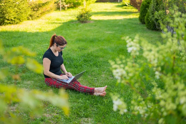 Une femme rousse est assise dans l'herbe verte d'un parc et fait du shopping en ligne. La fille maintient une distance sociale et travaille à l'extérieur sur un ordinateur portable. Un étudiant étudie à distance sur un ordinateur . — Photo