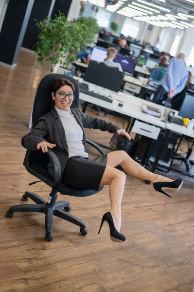 Mujer alegre en un traje monta en una silla de oficina en el lugar de trabajo mientras sus colegas están trabajando. Sonriente dama de negocios divirtiéndose durante el descanso en el trabajo en la oficina de espacio abierto. —  Fotos de Stock