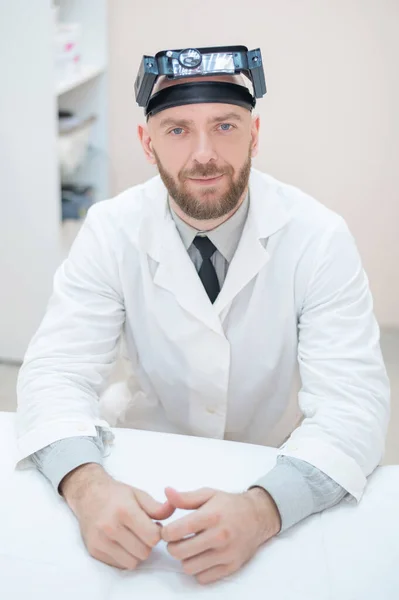 Maschio medico barbuto in un cappotto medico e con una cinghia di ingrandimento w luci seduto sulla scrivania. Attrezzature optometriche. Ufficio medico. — Foto Stock