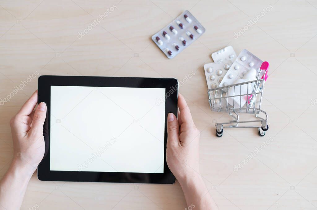 Digital tablet with a white screen in female hands and a mini cart with blisters of pills. The concept of a mobile application for the remote purchase of medicines. Online pharmacy.