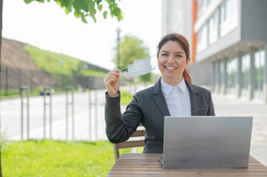 Mutlu iş kadını, yaz terasında dizüstü bilgisayar üzerinde çalışırken tıbbi maskesini çıkarıyor. Takım elbiseli gülümseyen bir kız kafede otururken özgürlüğün tadını çıkarıyor. Karantinanın sonu.