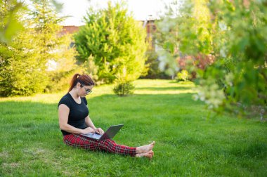 Kızıl saçlı bir kadın parktaki yeşil çimlerde oturuyor ve internetten alışveriş yapıyor. Kız sosyal mesafeyi koruyor ve dışarıda dizüstü bilgisayarla çalışıyor. Bir öğrenci uzaktan bilgisayarla çalışıyor..