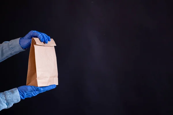 Weibliche Hände in einem blauen Handschuh halten eine braune Papiertüte vor schwarzem Hintergrund. Sichere Essenslieferung nach Hause. Ein Kurier im Jeanshemd reicht einem Kunden eine Bastelkartontasche. — Stockfoto