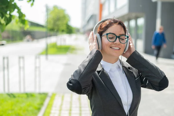 Retrato de una mujer sonriente en un traje de trabajo escuchando música en auriculares al aire libre. Empleada de oficina toma las manos en los auriculares disfrutando de la canción y bailando en la calle. —  Fotos de Stock