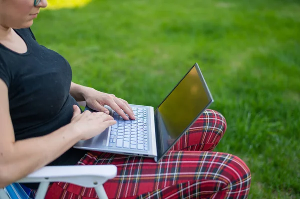 Kobieta siedzi w parku i typów na komputerze podczas zakupów online. Dziewczyna utrzymuje dystans społeczny i pracuje na zewnątrz na laptopie. Student uczy się zdalnie.. — Zdjęcie stockowe