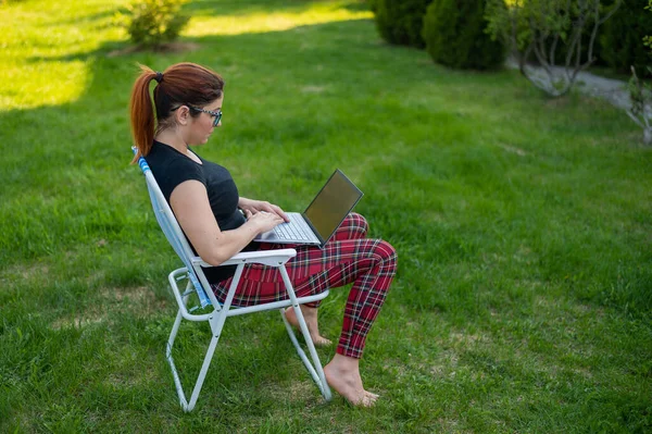 Uma mulher com óculos senta-se em uma cadeira dobrável e digita em um laptop. Freelancer feminino trabalha remotamente no jardim da casa de campo durante a quarentena. O trabalho é um prazer. . — Fotografia de Stock