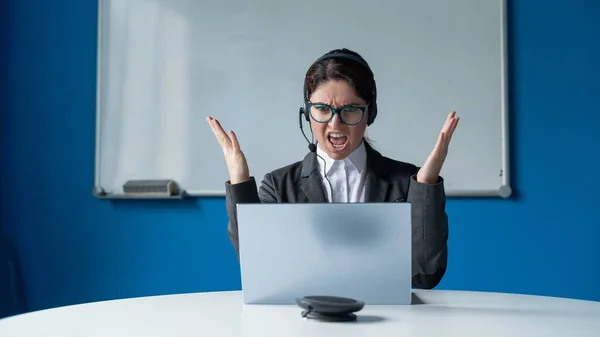En arg kvinna i ett headset har ett online-samtal på en bärbar dator i ett konferensrum. Kvinnlig chef som skällde ut underordnade för ett videosamtal. Framgångsrika förhandlingar på distans. — Stockfoto