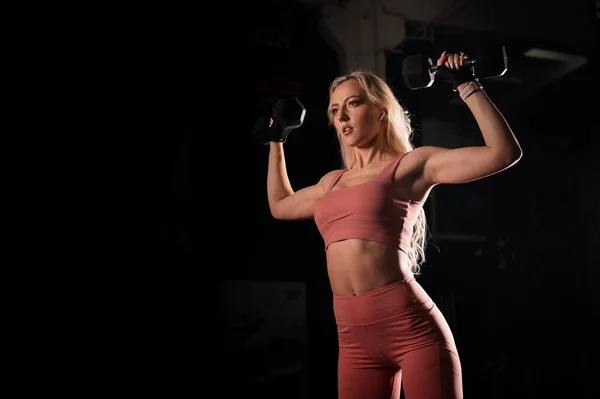 Ευρωπαία ξανθιά γυναίκα με αθλητική φιγούρα που φοράει γάντια κάνει ασκήσεις δικέφαλων με αλτήρα. Μοντέλο μπικίνι γυμναστικής που ποζάρει στο γυμναστήριο. Γυναίκα bodybuilder. — Φωτογραφία Αρχείου