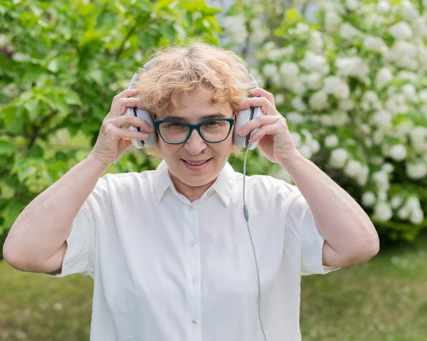 Щаслива стара жінка ходить в парку квітучих яблунь і одягає навушники. Жінка-пенсіонер насолоджується теплим літнім днем у саду і слухає музику. Передове старше покоління . — стокове фото