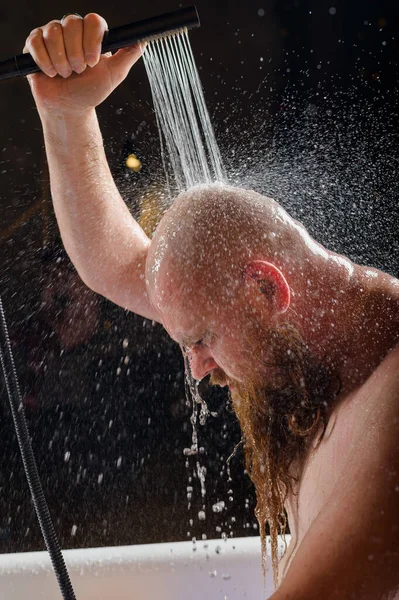 Il maschio si fa una doccia. Un uomo brutale con la barba rossa è in piedi nel bagno sotto l'acqua corrente e si lava. Spray scatter su uno sfondo nero. — Foto Stock