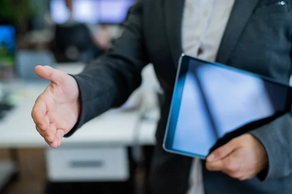 Business woman in a jacket and white shirt holding out a hand for a greeting. A faceless business woman holds a digital tablet and shakes her hand as an agreement on a successful transaction.