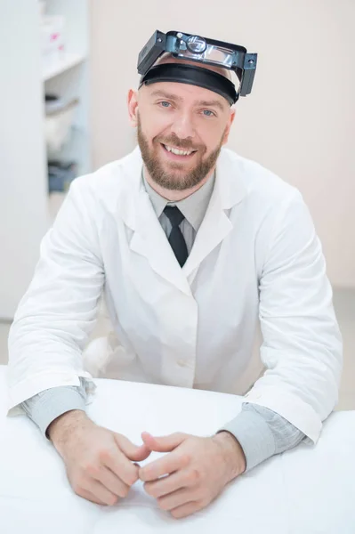 Mężczyzna brodaty lekarz w płaszczu medycznym i z powiększającym paskiem na głowie w świetle siedzącym na biurku. Sprzęt optyczny. Biuro medyczne. — Zdjęcie stockowe