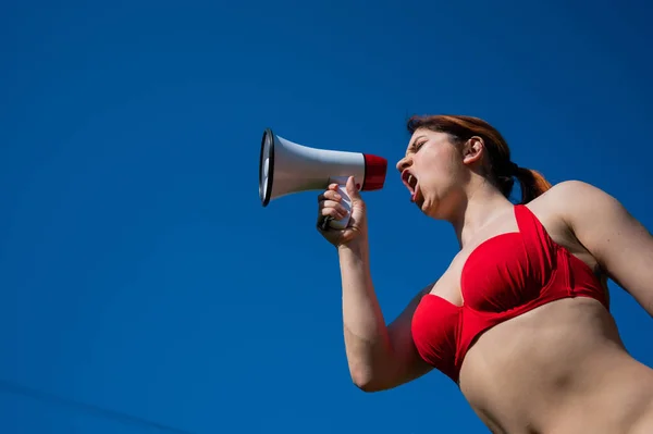 Femme blanche en bikini rouge travaille comme sauveteur sur la plage et crie à travers un mégaphone contre un ciel bleu. Une fille en maillot de bain est debout avec un haut-parleur. Vacances d'été. — Photo