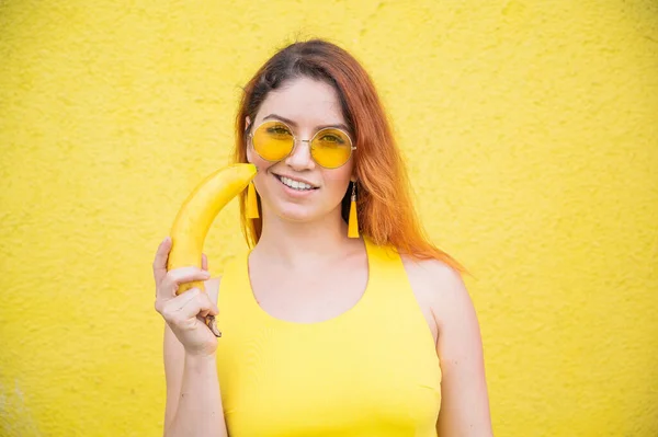 Porträtt av en vacker rödhårig kvinna i gul klänning och solglasögon håller en banan nära sensuella läppar. Flicka poserar med frukt på en gul bakgrund. — Stockfoto