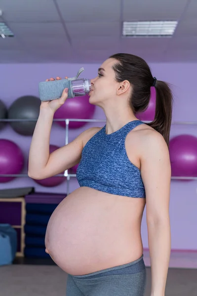 Беременная женщина в спортивном костюме пьет воду в тренажерном зале с фитнес-мечами. Девушка занимается пилатесом для будущих мам. Последний триместр . — стоковое фото